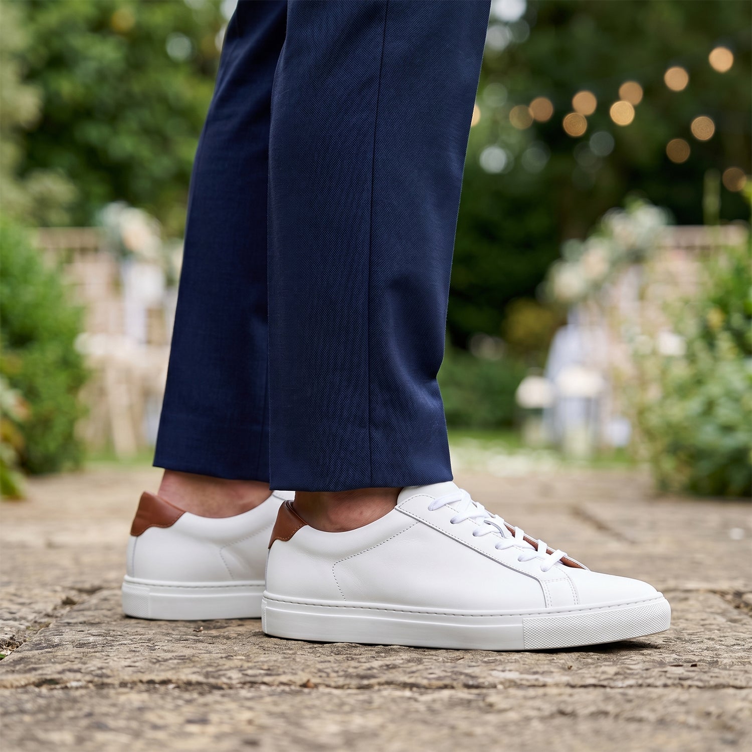 Close-up of a man's legs wearing tailored navy blue suit trousers with a sharp no-break hem, perfectly paired with immaculate white premium leather trainers, set against a softly blurred outdoor wedding background.