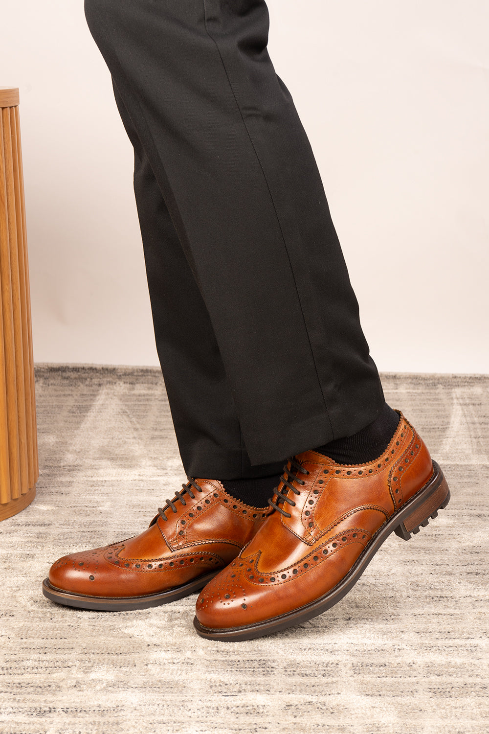 Brown dress shoes worn with black pants on a light wooden floor.