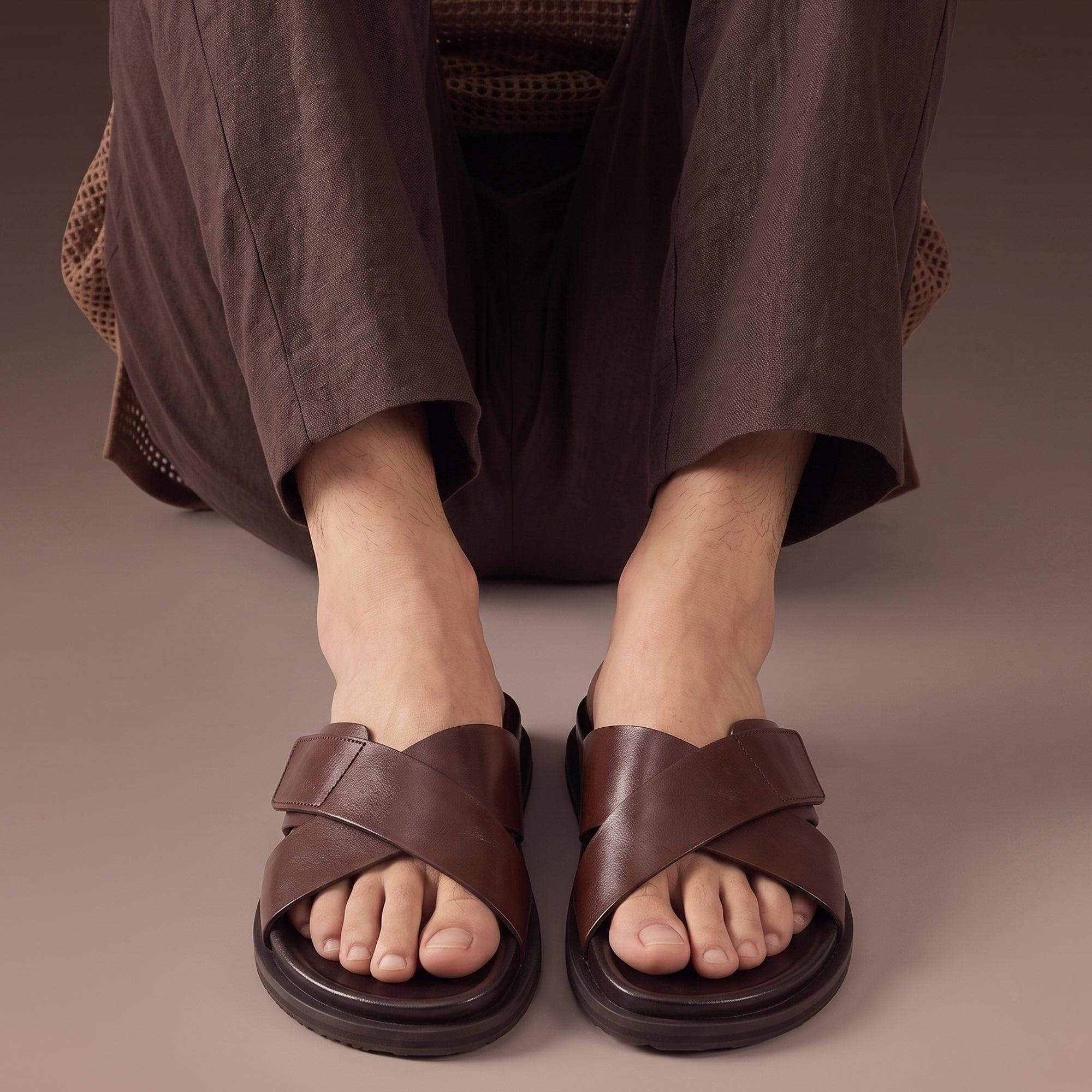 Men's dark brown leather cross-strap slide sandals worn barefoot with casual brown summer trousers.