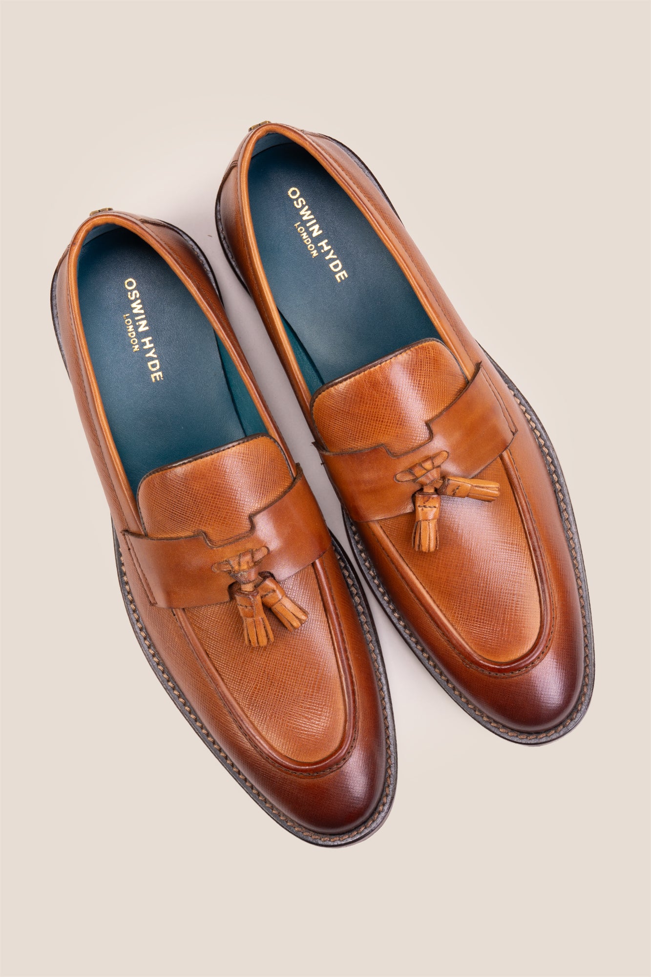 Premium tan leather tassel loafers for men featuring a textured finish, burnished moc-toe details, decorative leather tassels, and a cushioned teal interior.