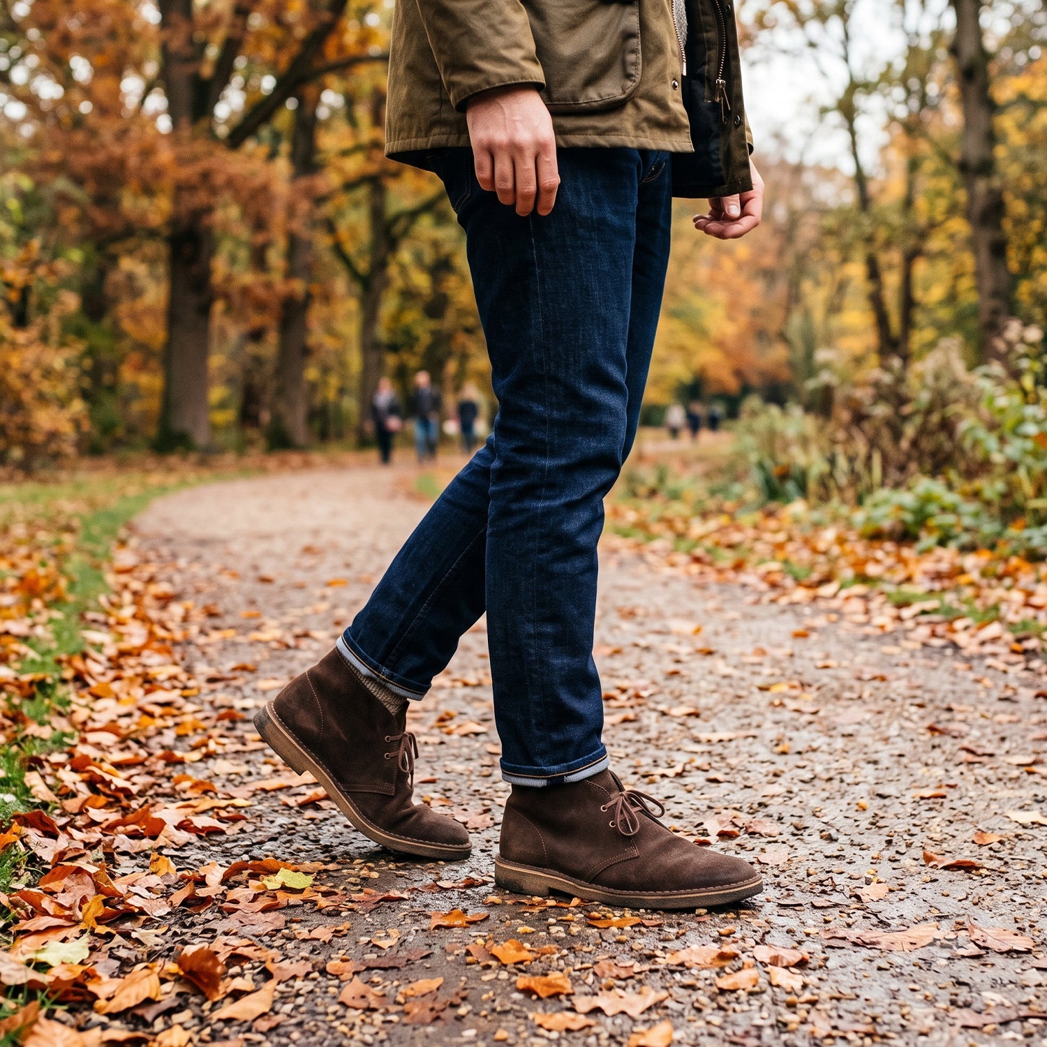 Man wearing dark blue slim-fit jeans and brown suede chukka boots walking on a park path covered in fallen autumn leaves.