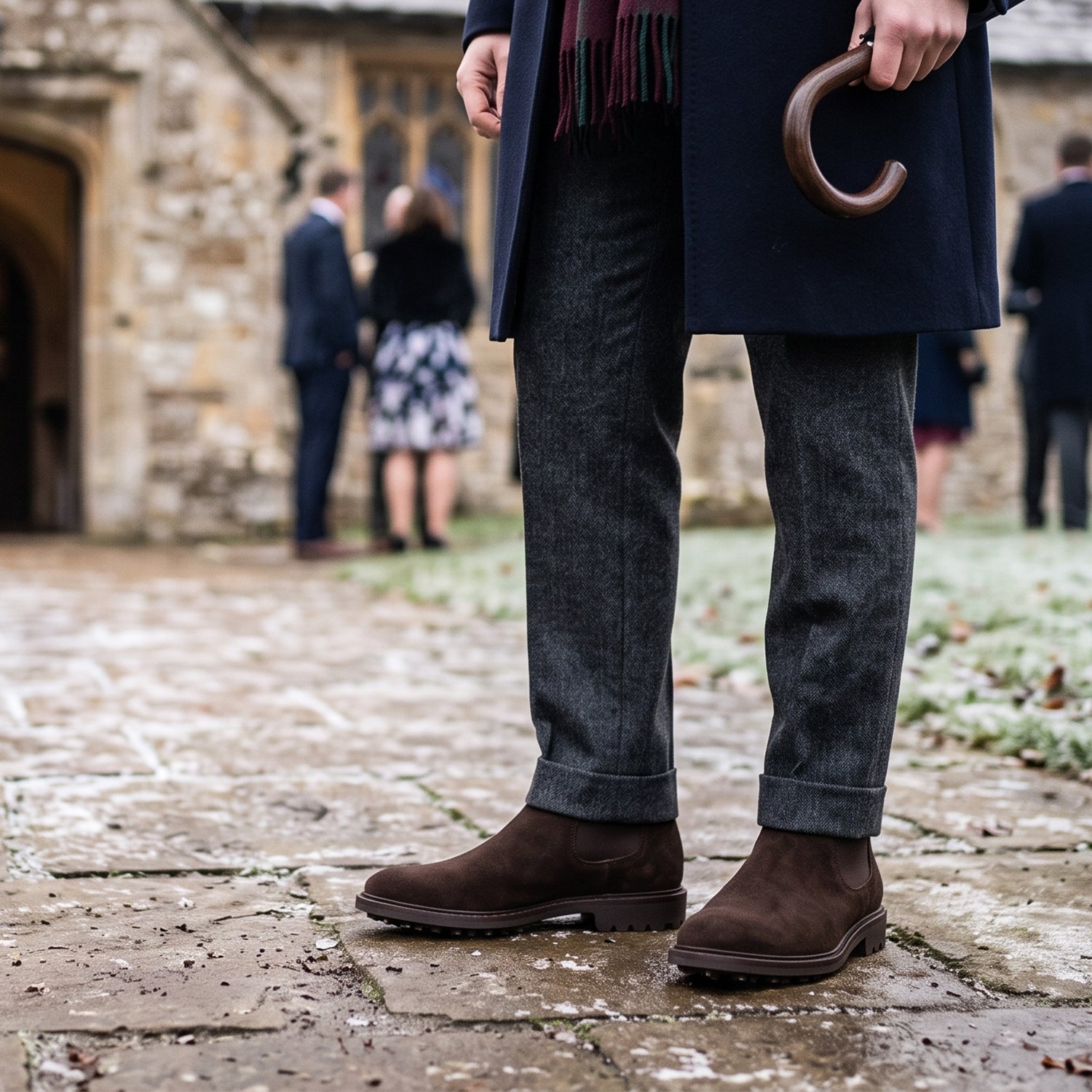 Close-up detail of a pair of rich brown suede Chelsea boots and the polished dark wood handle of an umbrella