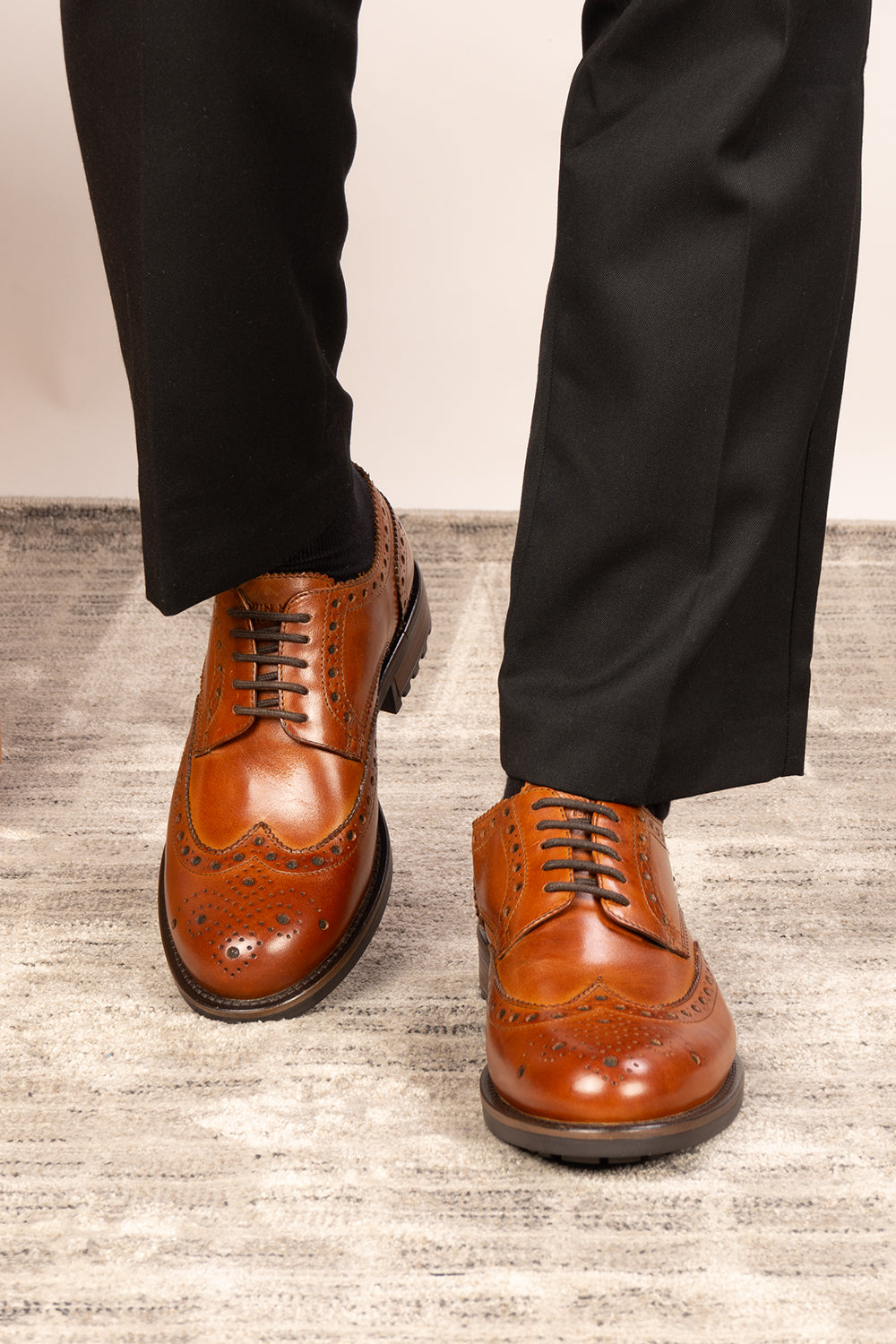 Brown leather dress shoes worn with black pants on a light wooden floor.
