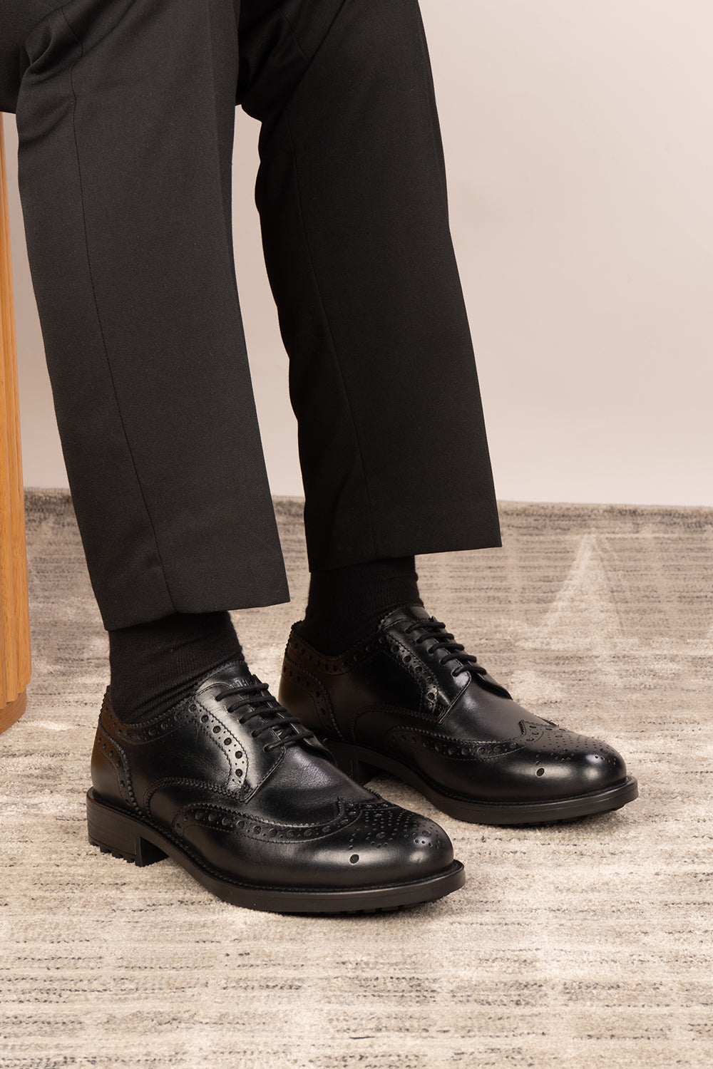 Black dress shoes worn with black pants on a wooden floor.