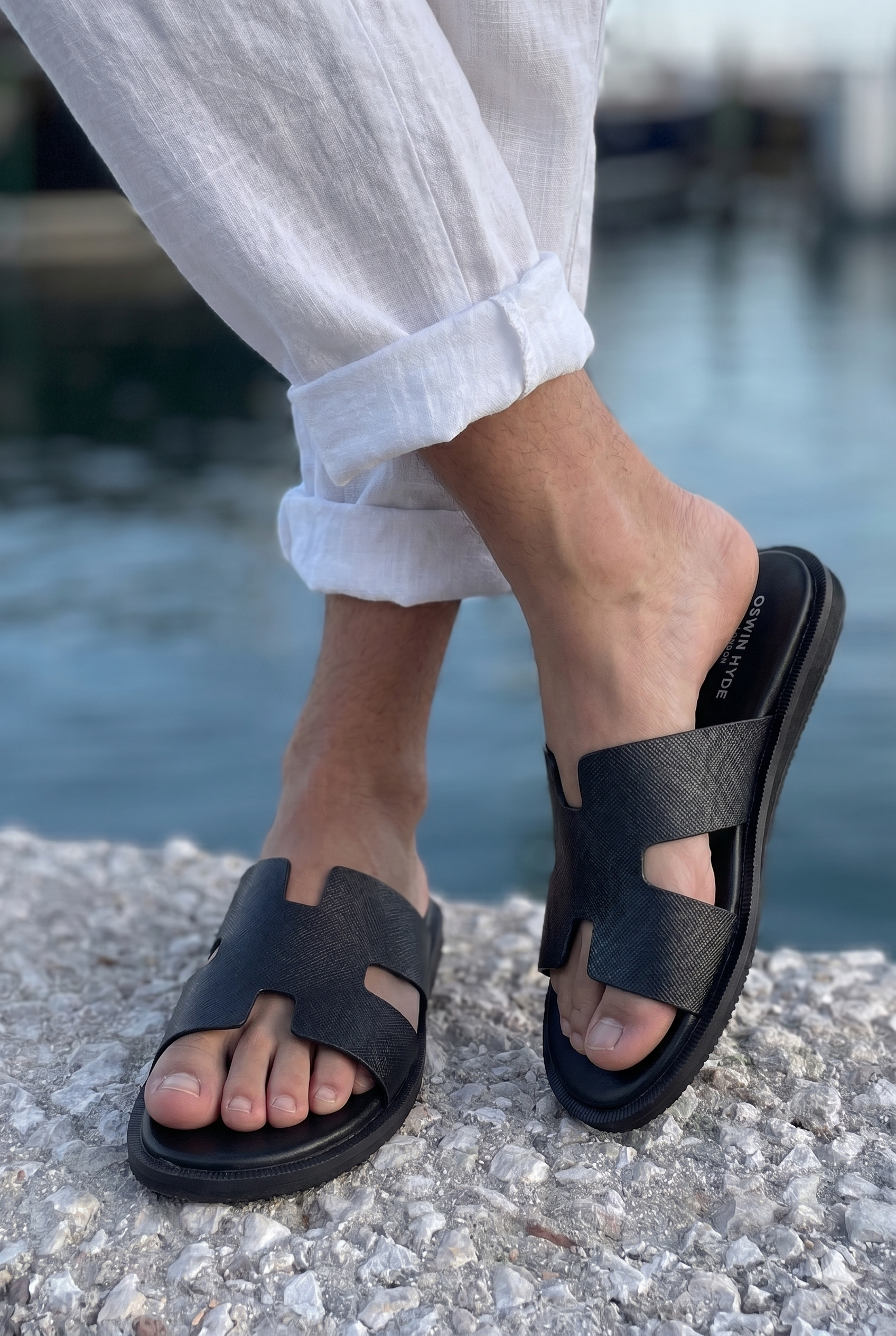Model wearing Men’s Phoenix Black sandals in premium Saffiano leather featuring the signature hyde strap H-frame design.