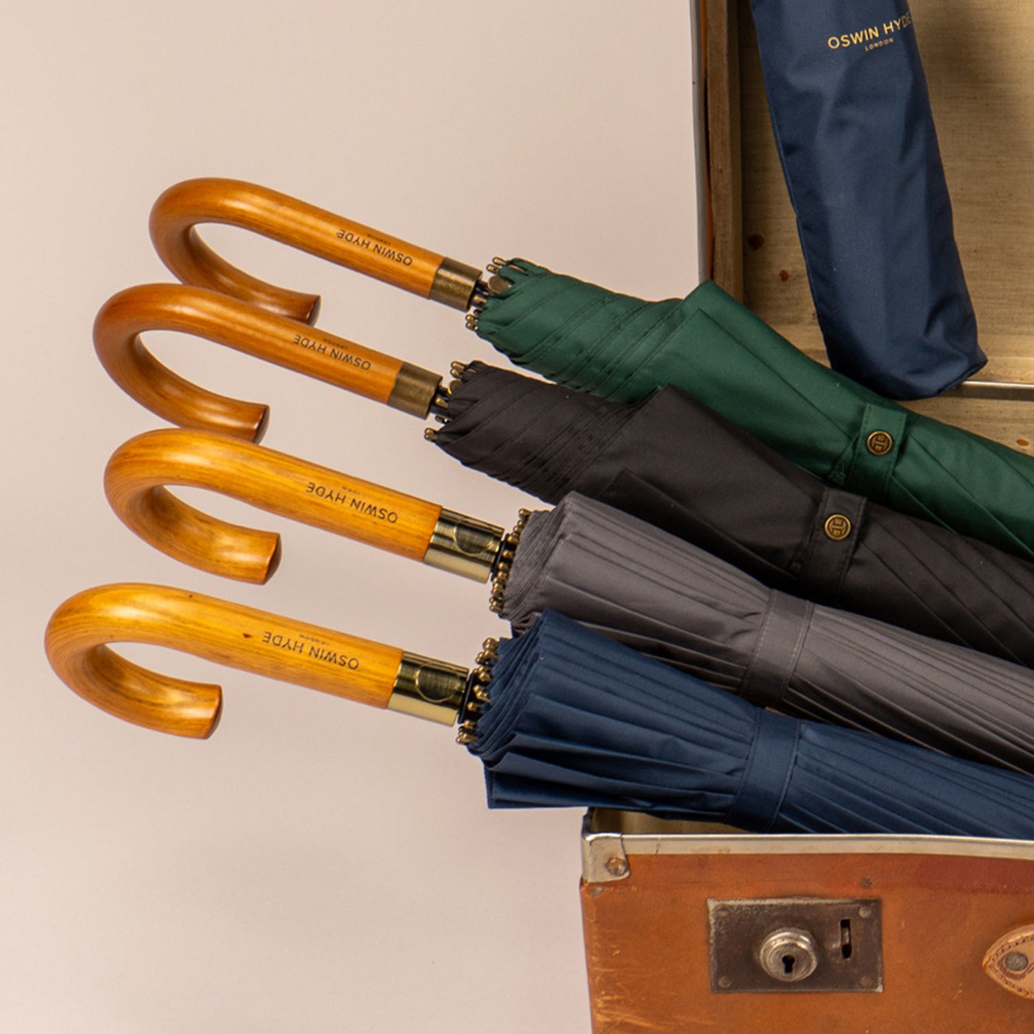 Premium Oswin Hyde London umbrellas in green, black, grey, and navy blue neatly folded inside a vintage leather suitcase. The umbrellas feature classic J-shaped wooden handles with brass accents and buttoned closure straps.