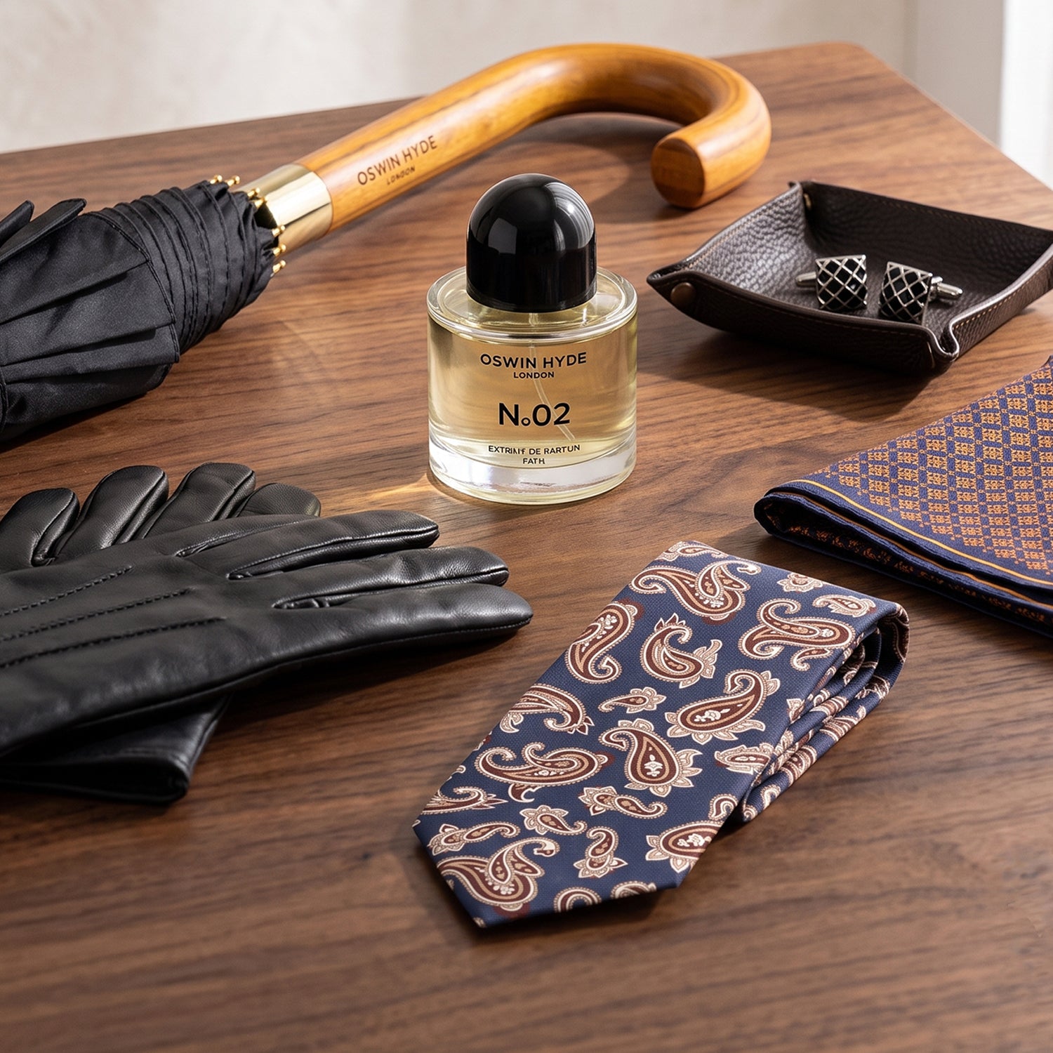 Luxury men's accessories collection by Oswin Hyde London including leather gloves, a navy paisley silk tie, a geometric pattern pocket square, silver cufflinks in a tray, a classic umbrella, and No.02 Extrait de Parfum on a wooden background.