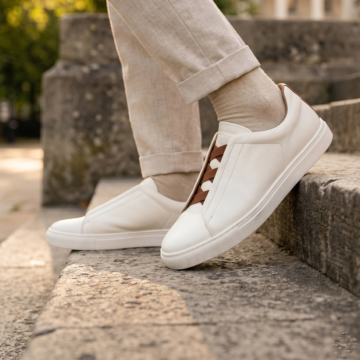 Premium men's white leather smart casual trainers featuring a laceless design with brown elasticated cross straps, matching brown heel tab, and white rubber cupsole.