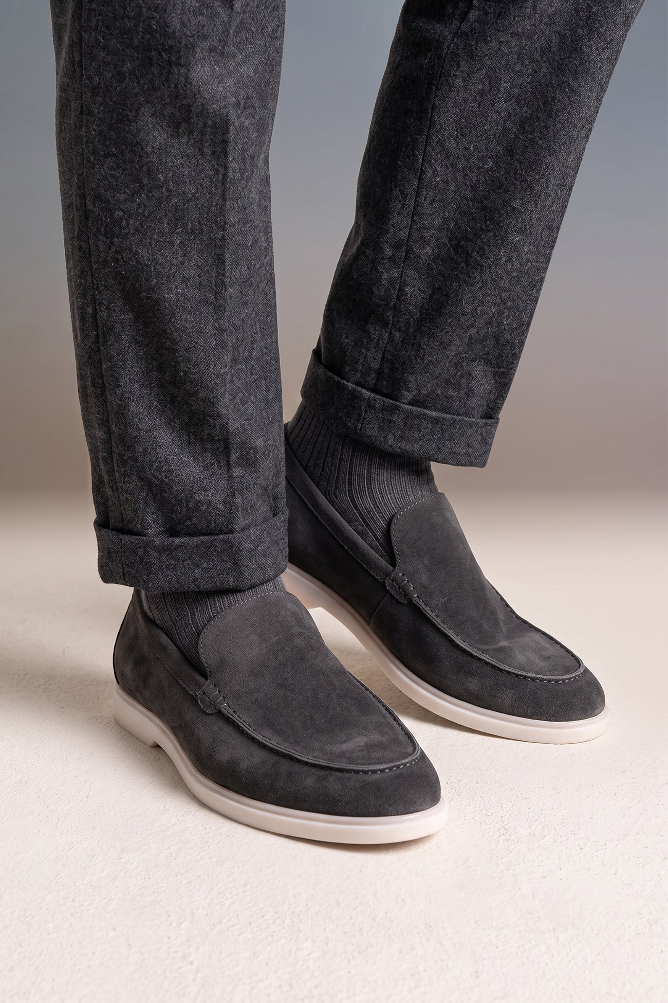 A person wearing Oswin Hyde Morgan grey suede loafers with charcoal grey trousers and ribbed grey socks.