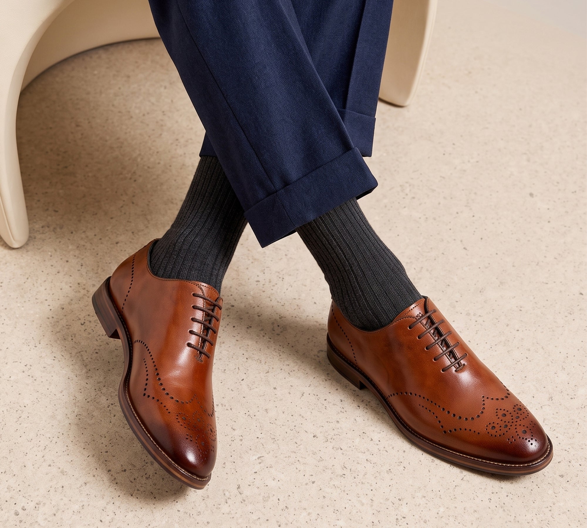 Men's tan leather wingtip brogue oxfords featuring classic perforation, worn with dark grey dress socks and navy suit pants.