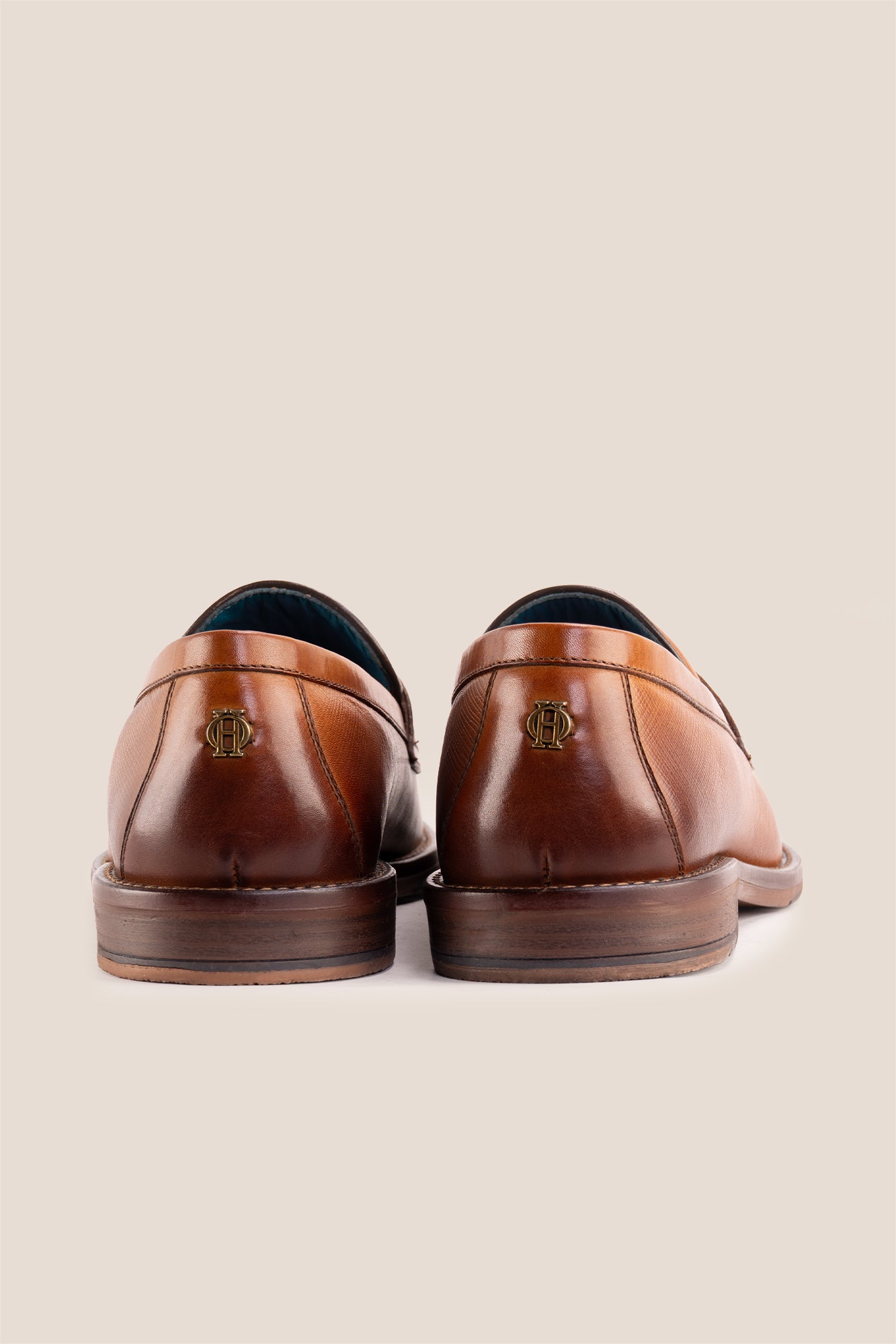 Premium tan leather tassel loafers for men featuring a textured finish, burnished moc-toe details, decorative leather tassels, and a cushioned teal interior.