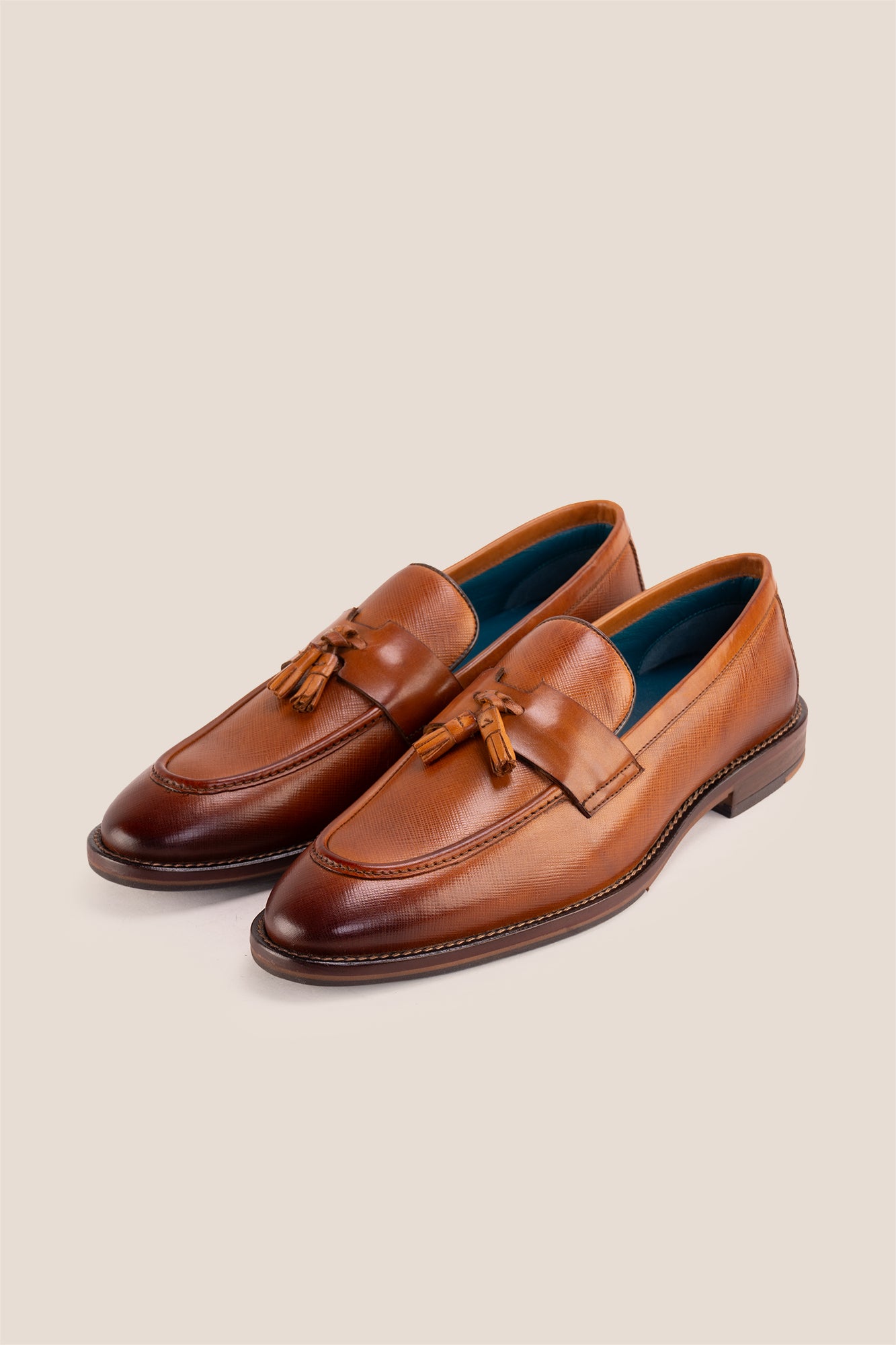 Premium tan leather tassel loafers for men featuring a textured finish, burnished moc-toe details, decorative leather tassels, and a cushioned teal interior.