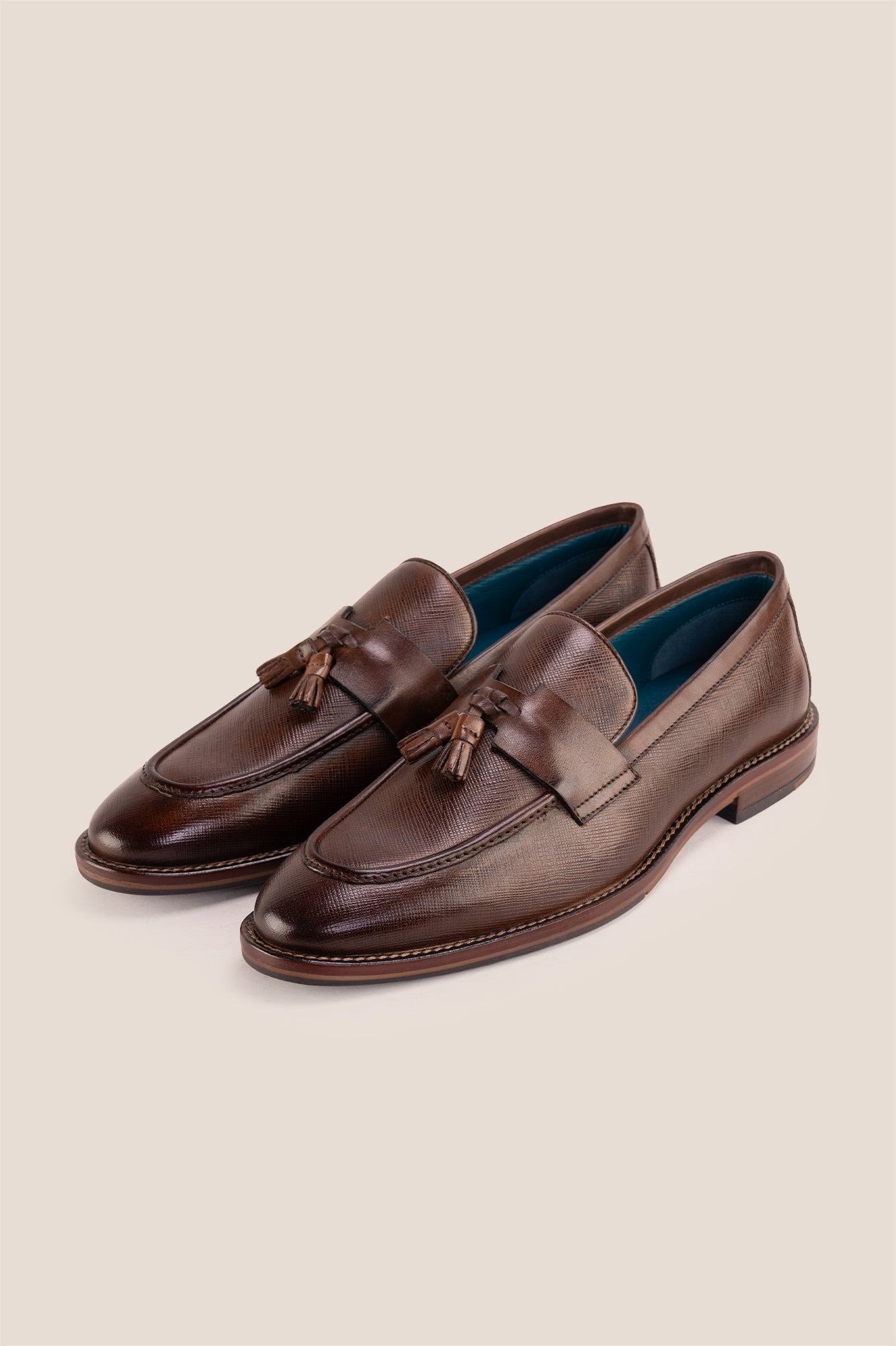 Premium brown leather tassel loafers for men featuring a textured finish, burnished moc-toe details, decorative leather tassels, and a cushioned teal interior.